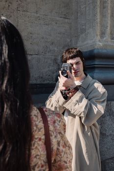 Man in a trench coat photographing a woman with an analog camera outdoors.