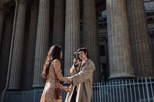 A couple holding hands while filming outside a historic columned building, capturing a romantic moment.
