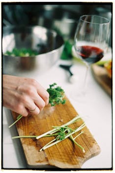 A hand prepares fresh herbs on a wooden cutting board beside a glass of red wine in a kitchen setting.