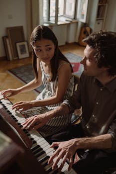 A man and woman sitting side by side playing piano in a cozy indoor setting with natural light.