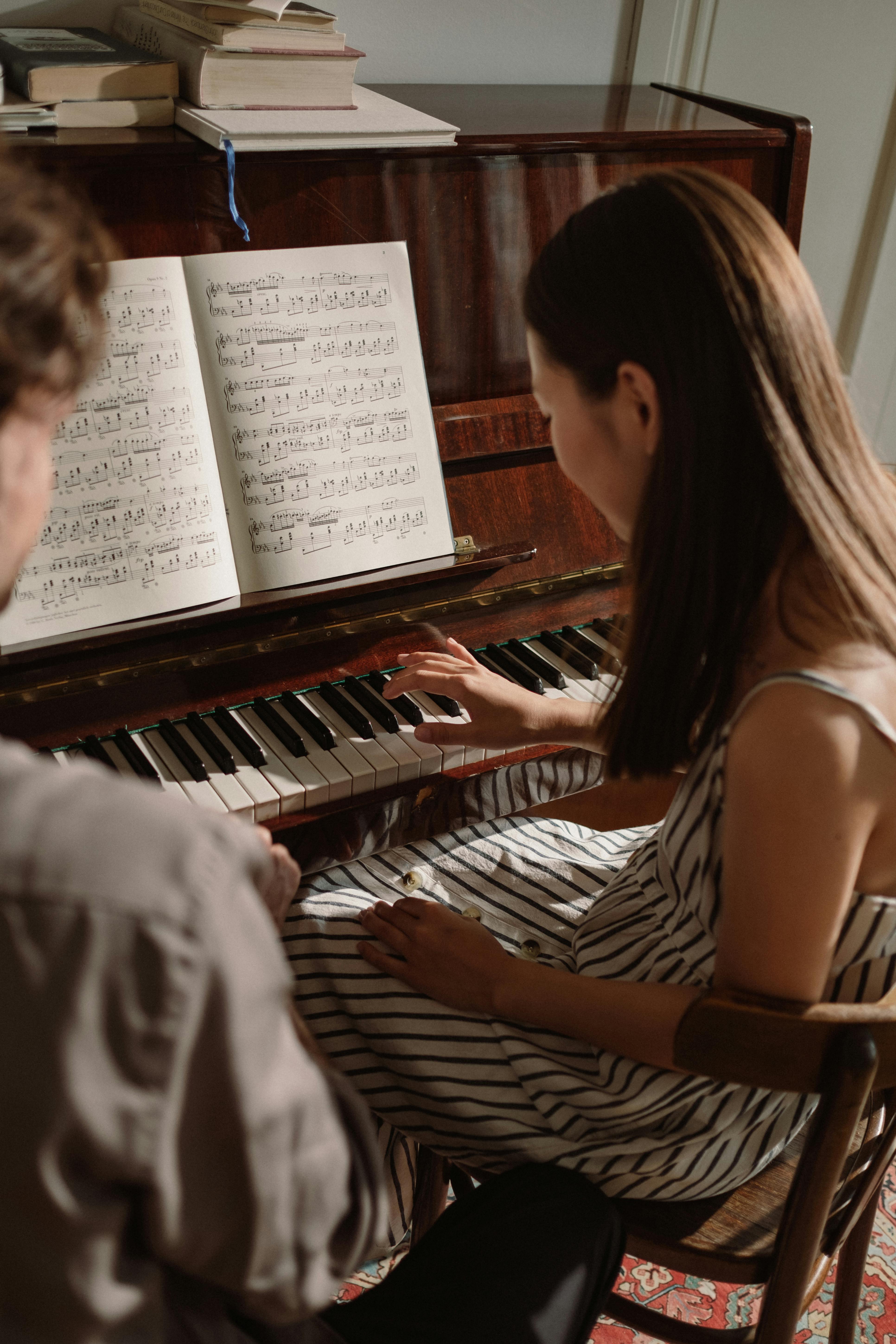 Woman in striped dress plays piano indoors with music sheets, accompanied by another person.