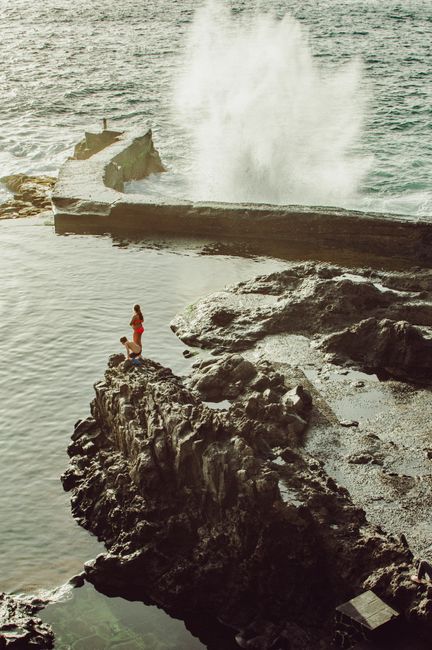 A person stands on a rocky cliff in Tenerife, waves crashing dramatically.