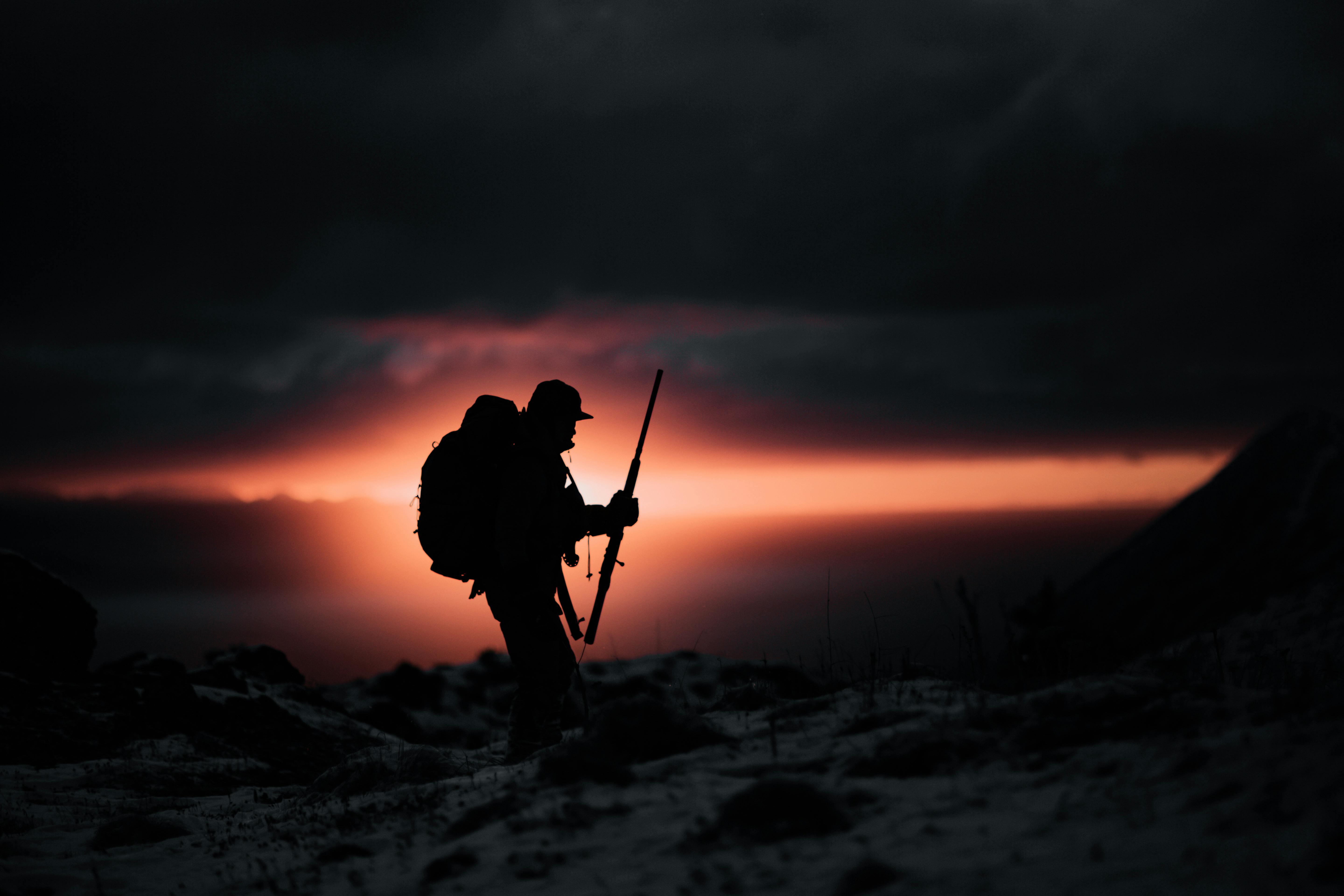 Dramatic silhouette of a hunter with rifle against a fiery twilight sky in rugged mountain terrain.