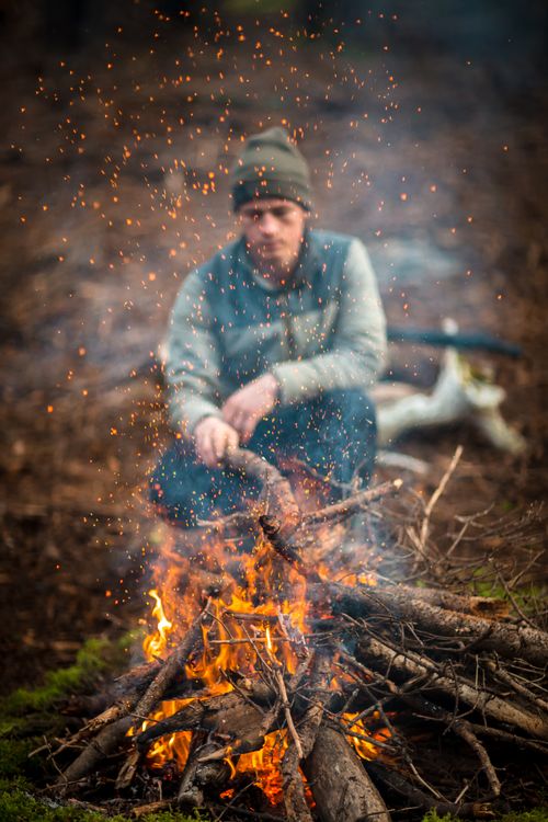 Free A man crouches near a blazing campfire in a forest setting, surrounded by sparks. Stock Photo