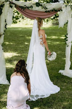Bride in wedding gown with veil stands under floral arch outdoors, holding bouquet.
