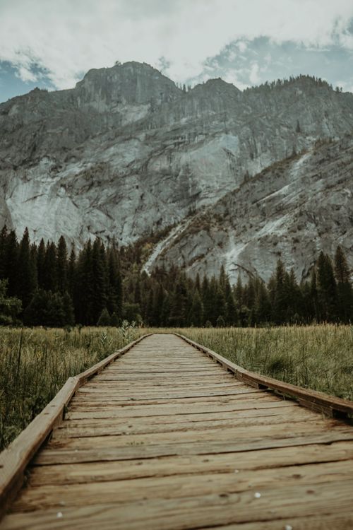 Free Discover tranquility on a wooden boardwalk leading to towering mountain peaks under a cloudy sky. Stock Photo