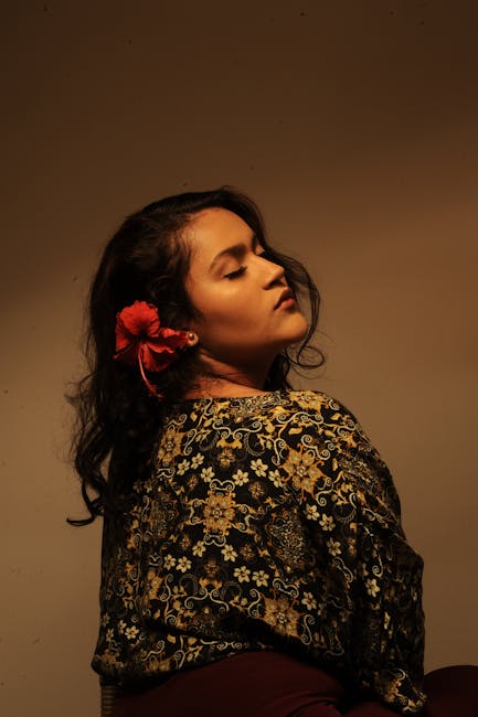 Portrait of a woman with closed eyes and floral hair accessory against a dark background.