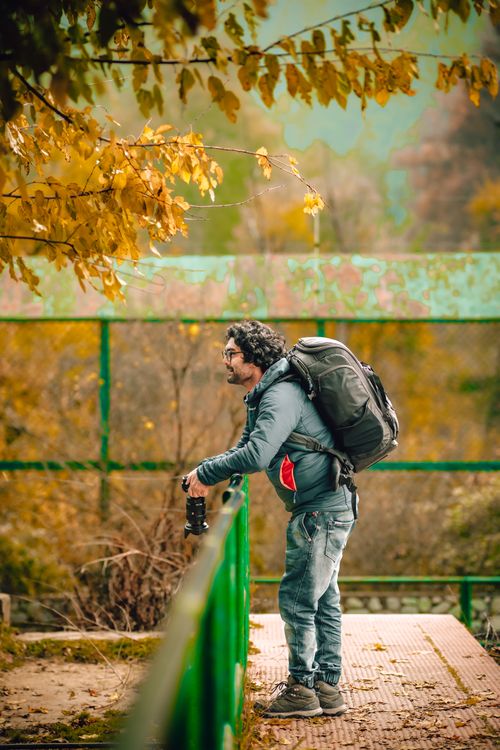 Free Man with camera and backpack gazes at fall foliage in a serene park setting. Stock Photo