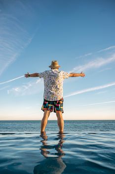 A man enjoys a scenic view of the ocean, standing by the poolside with arms outstretched and a fedora hat under a clear blue sky.
