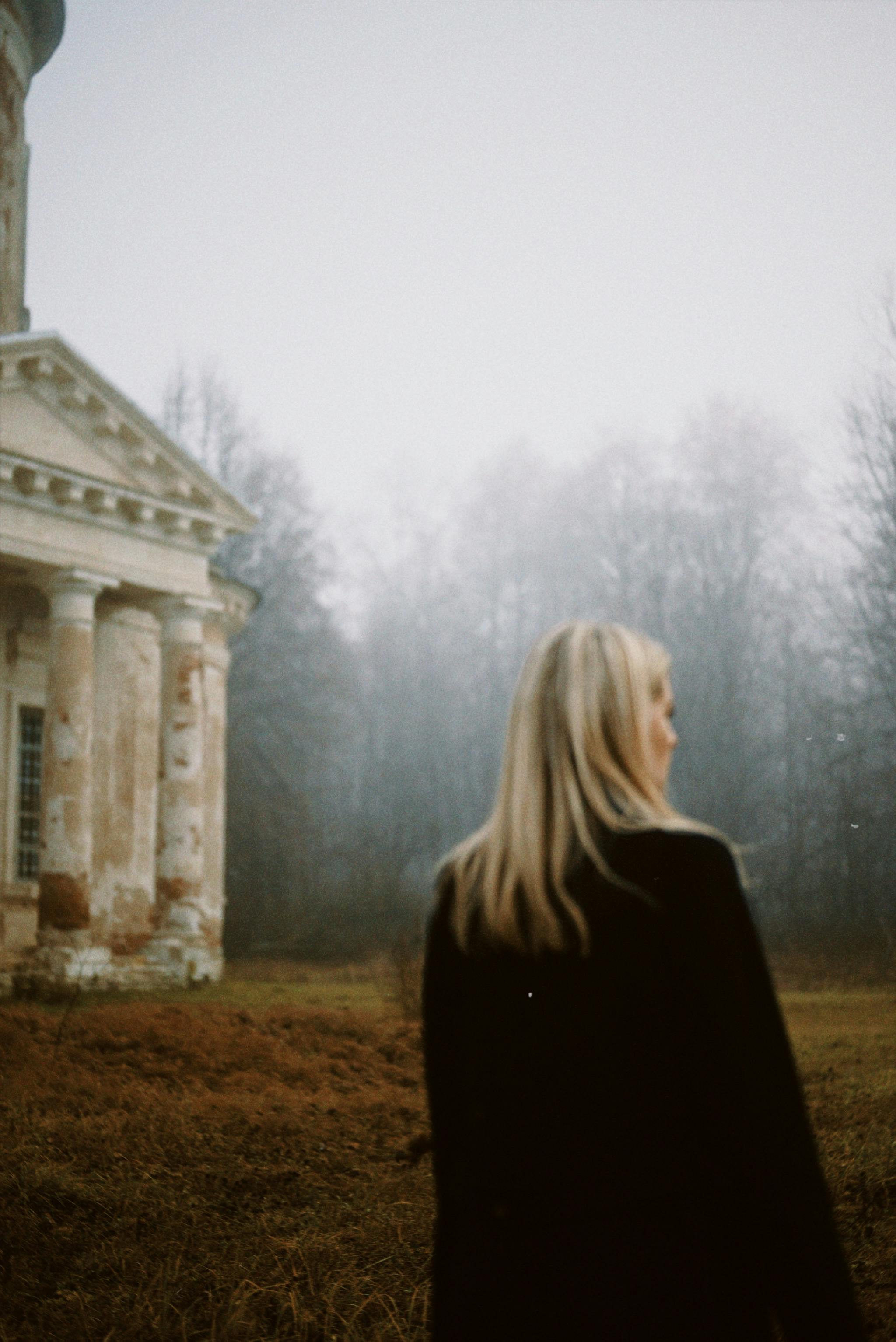 A woman in a black coat walks past an abandoned building amidst fog and a forest setting.