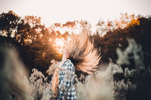 A woman with tousled hair flips her head back in a rural meadow during sunset.