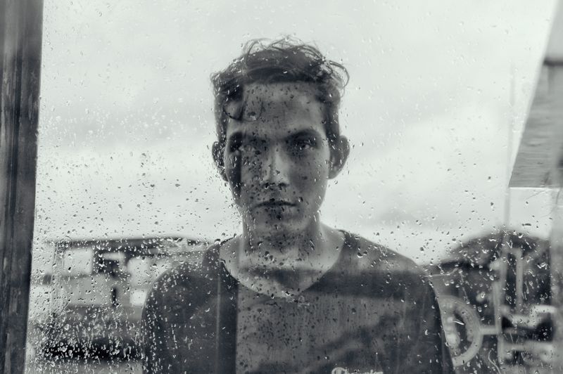 Moody black and white portrait of a man behind a rain-soaked window, exuding introspection.