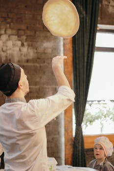 A chef skillfully tosses pizza dough in a cozy kitchen setting, observed by an intrigued child.