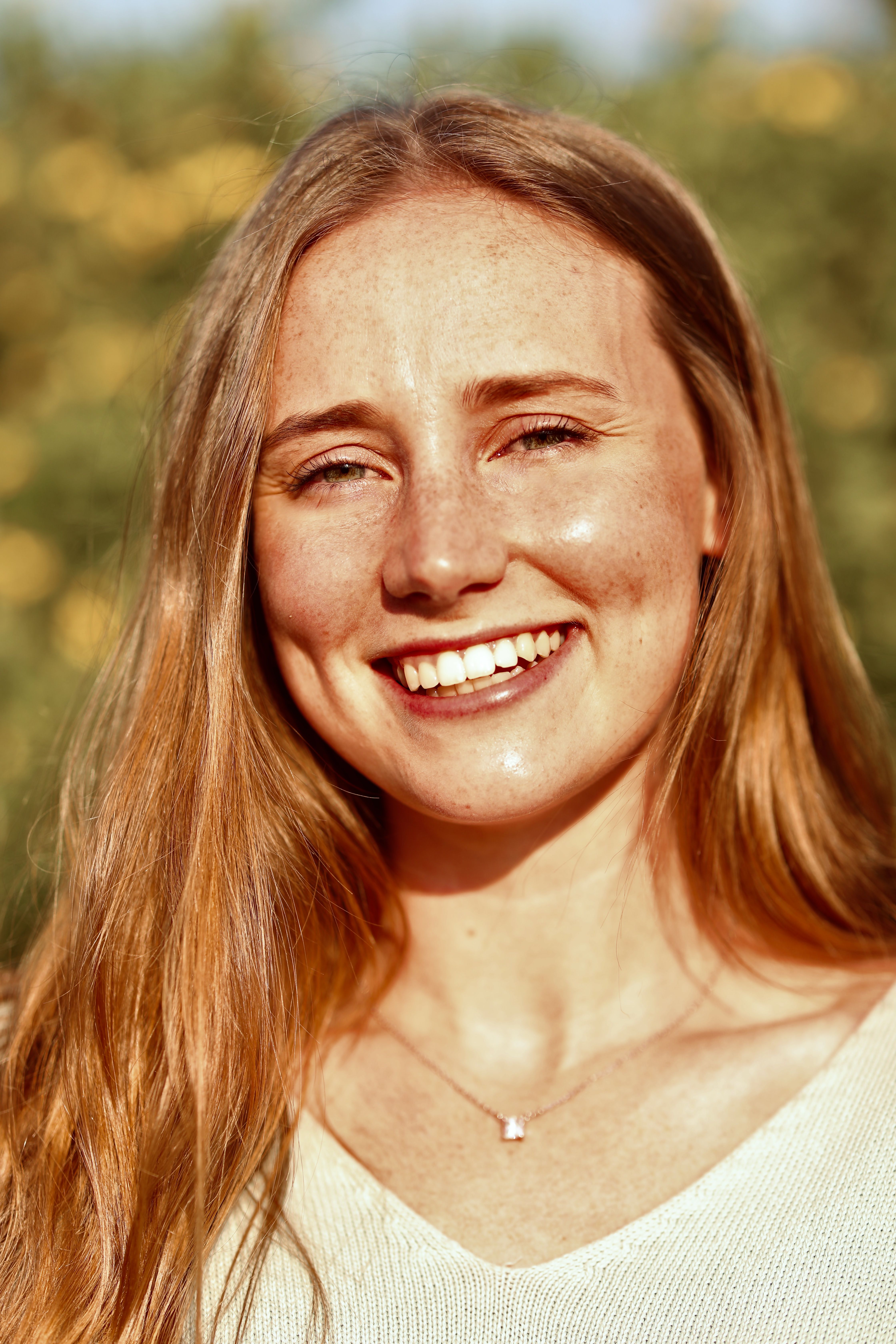 A cheerful woman with long hair smiles warmly in a sunny outdoor setting.