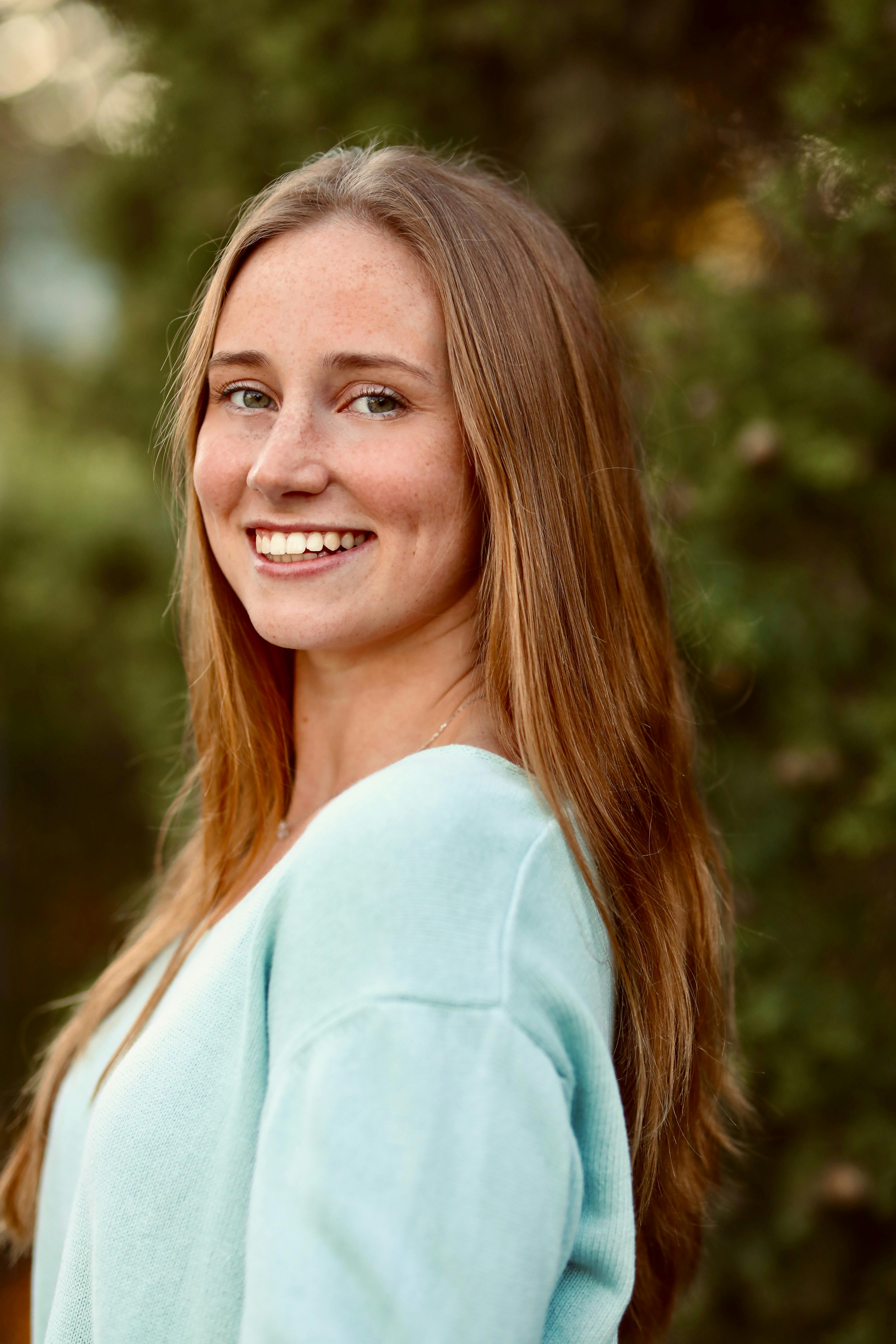 A cheerful young woman with long hair smiles in an outdoor setting, wearing a light blue sweater.