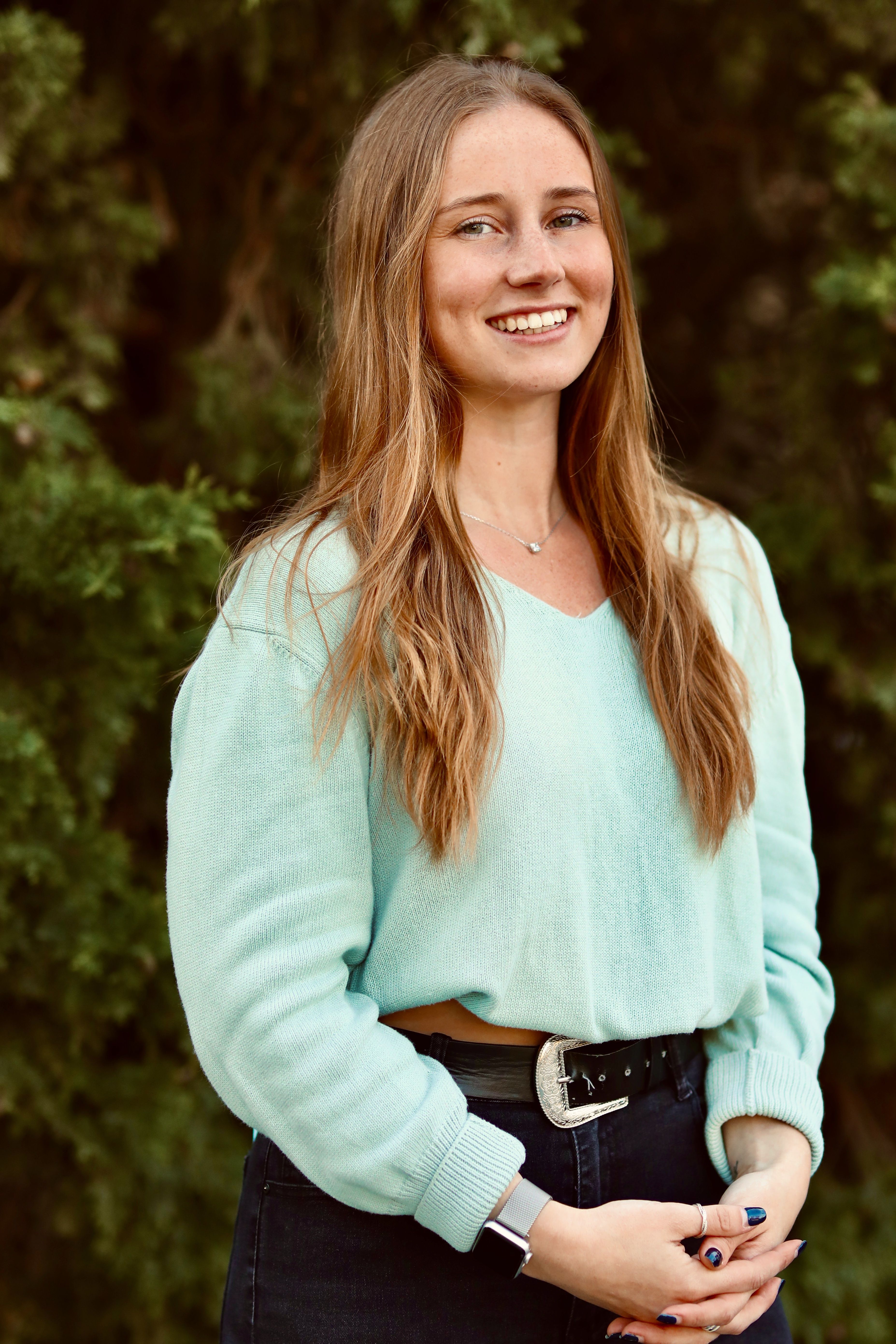 Smiling woman with long blond hair in a blue top standing outdoors against a green background.