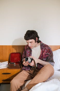 A young man with vintage style sits on a bed and examines a classic camera.