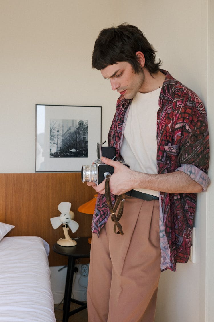 Young Man Leaning Against Wall In Bedroom And Holding Analog Camera