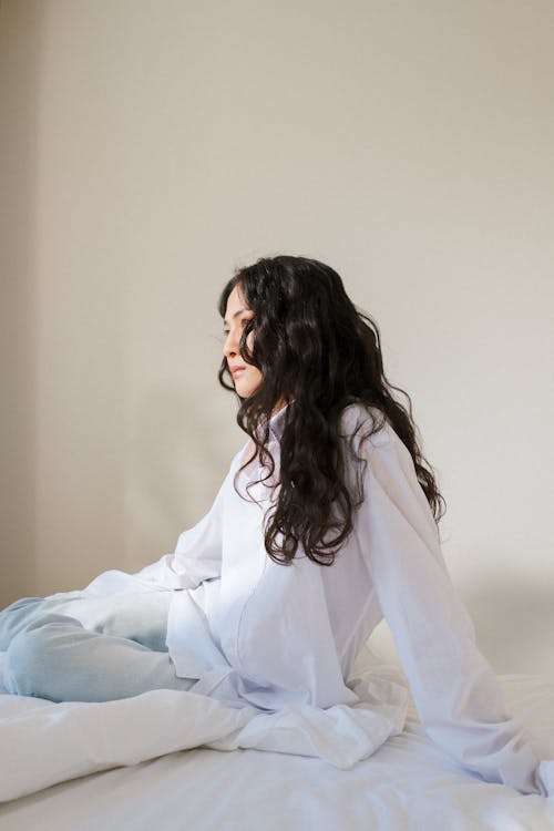 Free Young Woman with Black Curly Hair Sitting in Bed in White Pyjamas Stock Photo