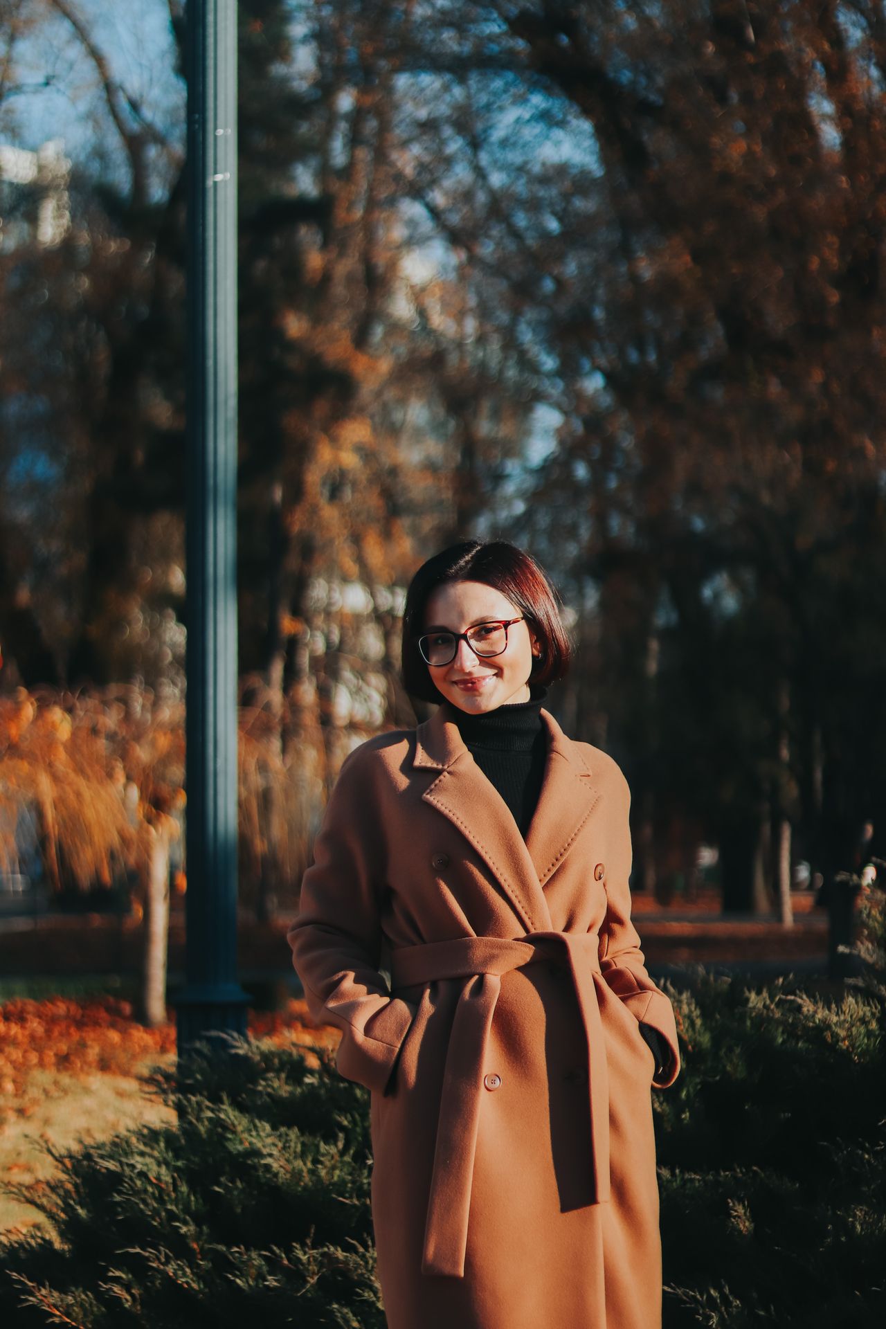 Woman wearing a rust-brown belted trench coat standing in an autumn park