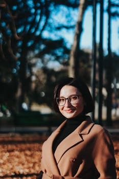 A woman in a brown coat and glasses poses confidently in an outdoor setting with blurred trees.
