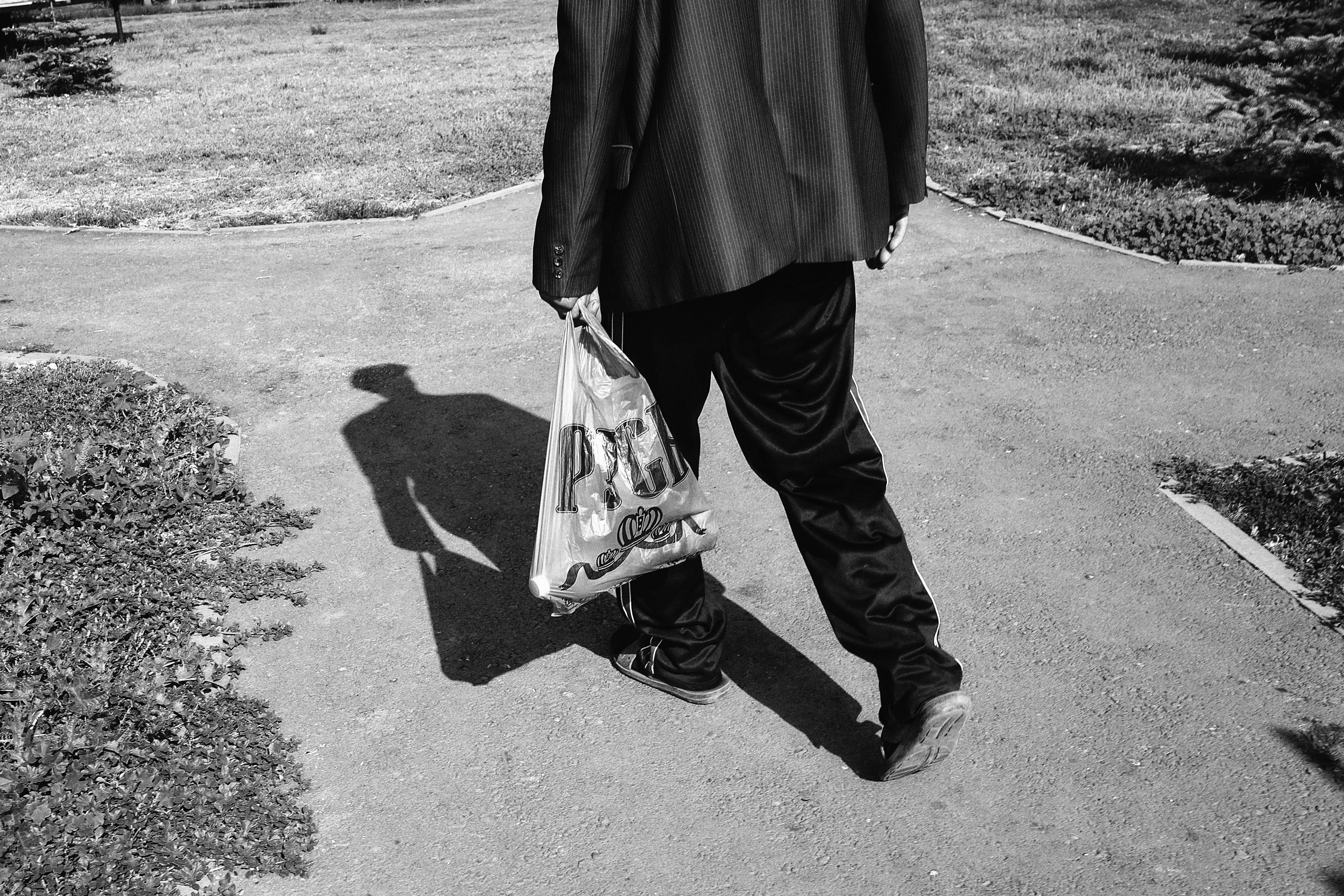 A black and white image capturing a man's back view as he walks on a sidewalk holding a bag.