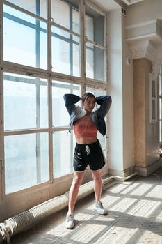 A woman in activewear poses confidently by a large window in a sunlit room.