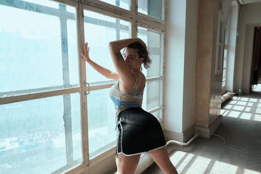 A woman poses confidently by a large sunlit window, showcasing style and tattoo art.