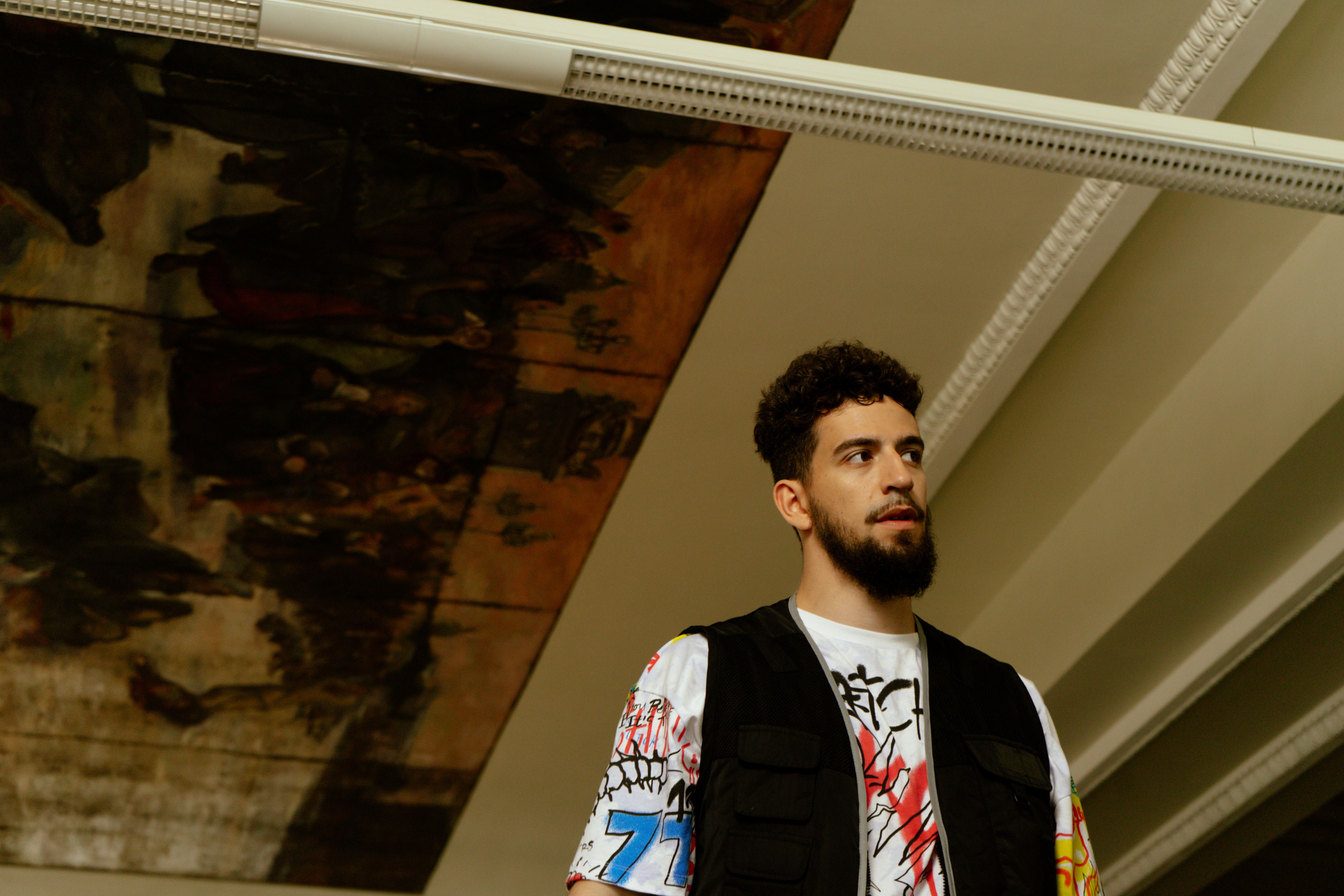A bearded man wearing a printed vest stands indoors beneath an artistic ceiling.
