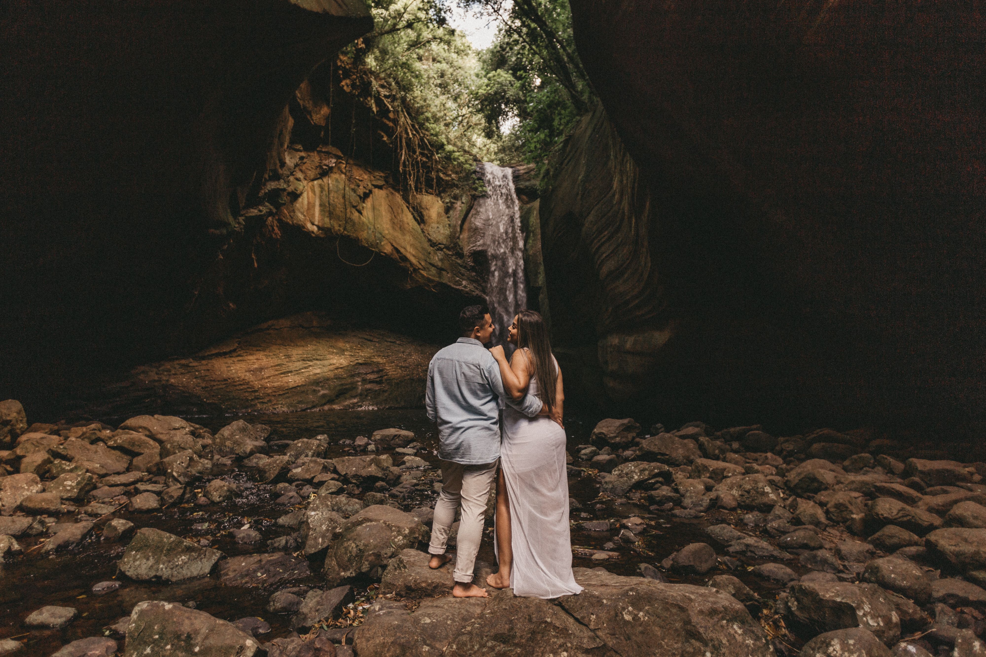 A couple stands embracing near a waterfall inside a rocky cave, surrounded by nature.