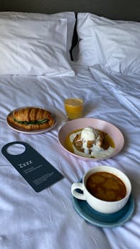 Indulge in a cozy breakfast in bed featuring croissants, coffee, and juice on white linen.