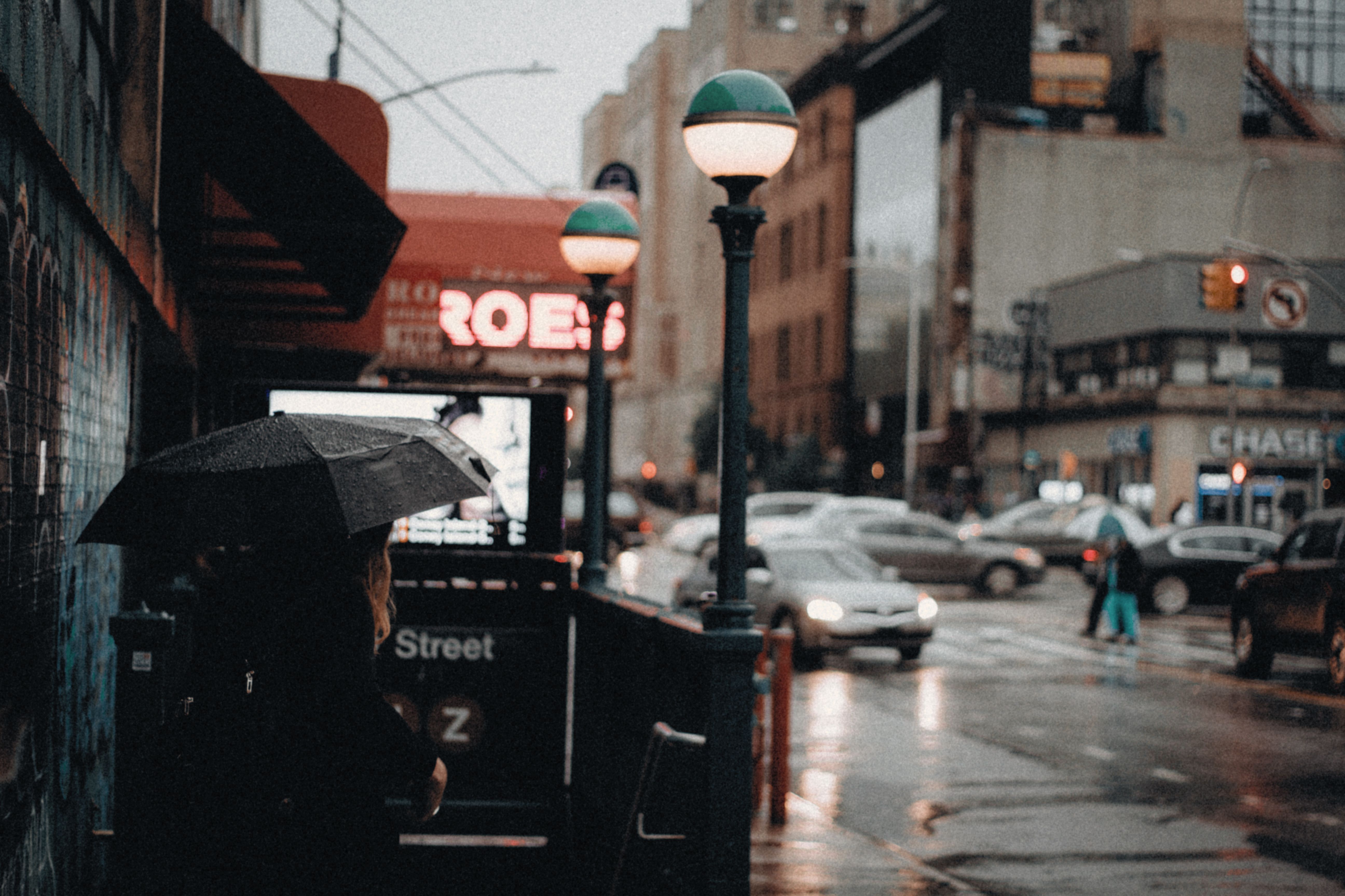 A rainy city street with a person holding an umbrella by a busy intersection.