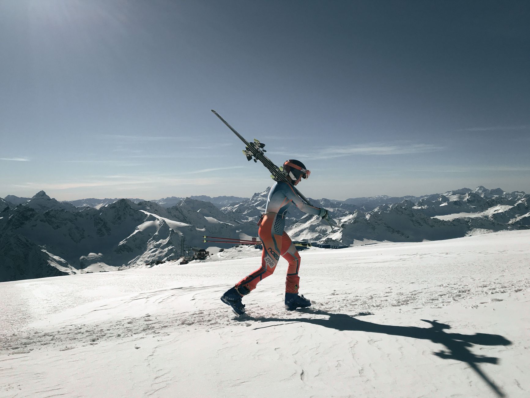 A skier carrying skis on a snowy mountain with a panoramic view of the Alps under a clear sky.
