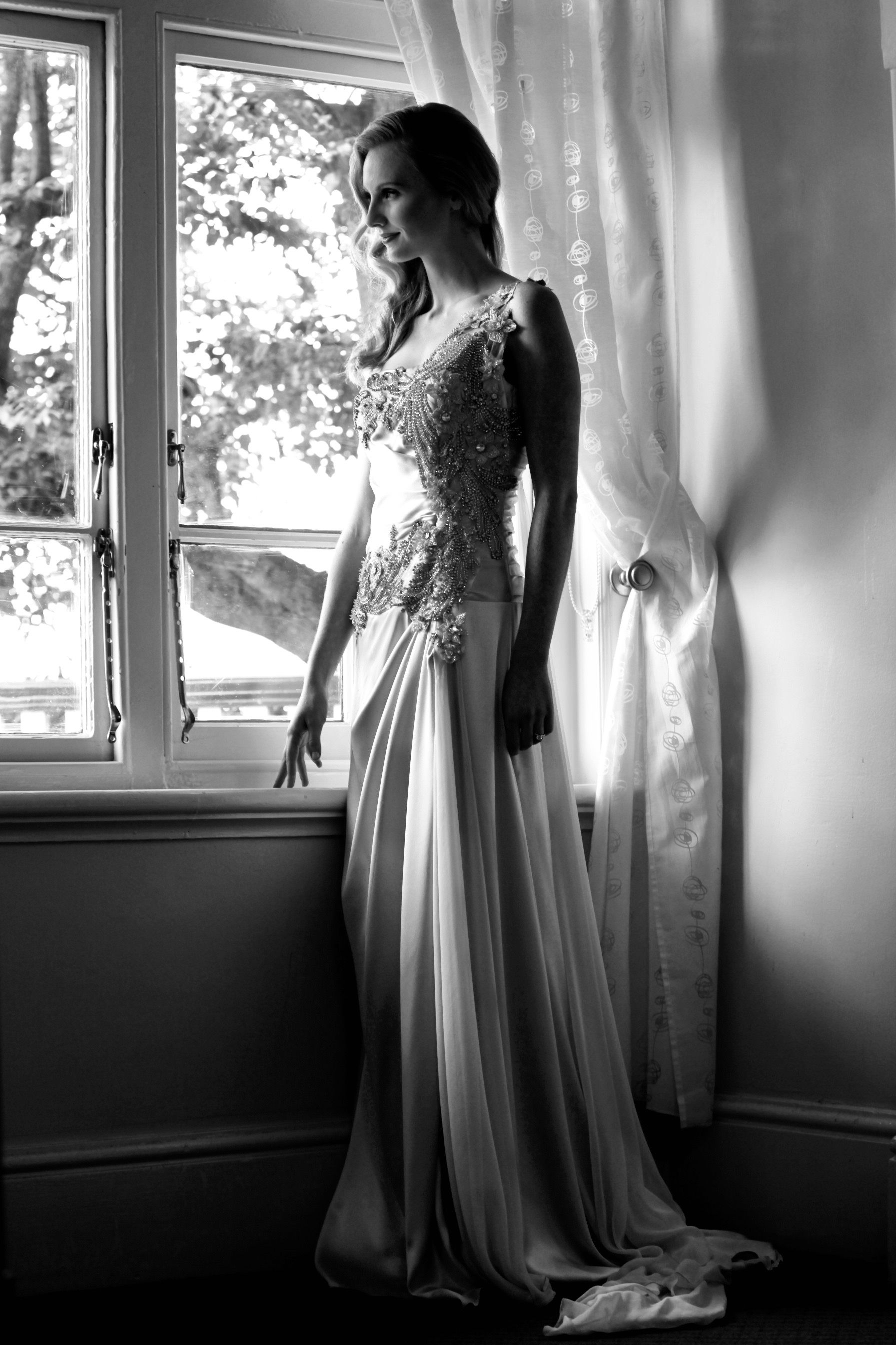 A bride in an ornate gown stands by a window in a black and white portrait.
