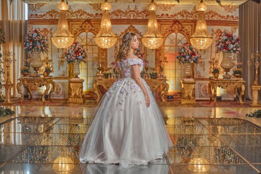 Woman in ornate floral gown stands in a grand setting with chandeliers and flower arrangements.