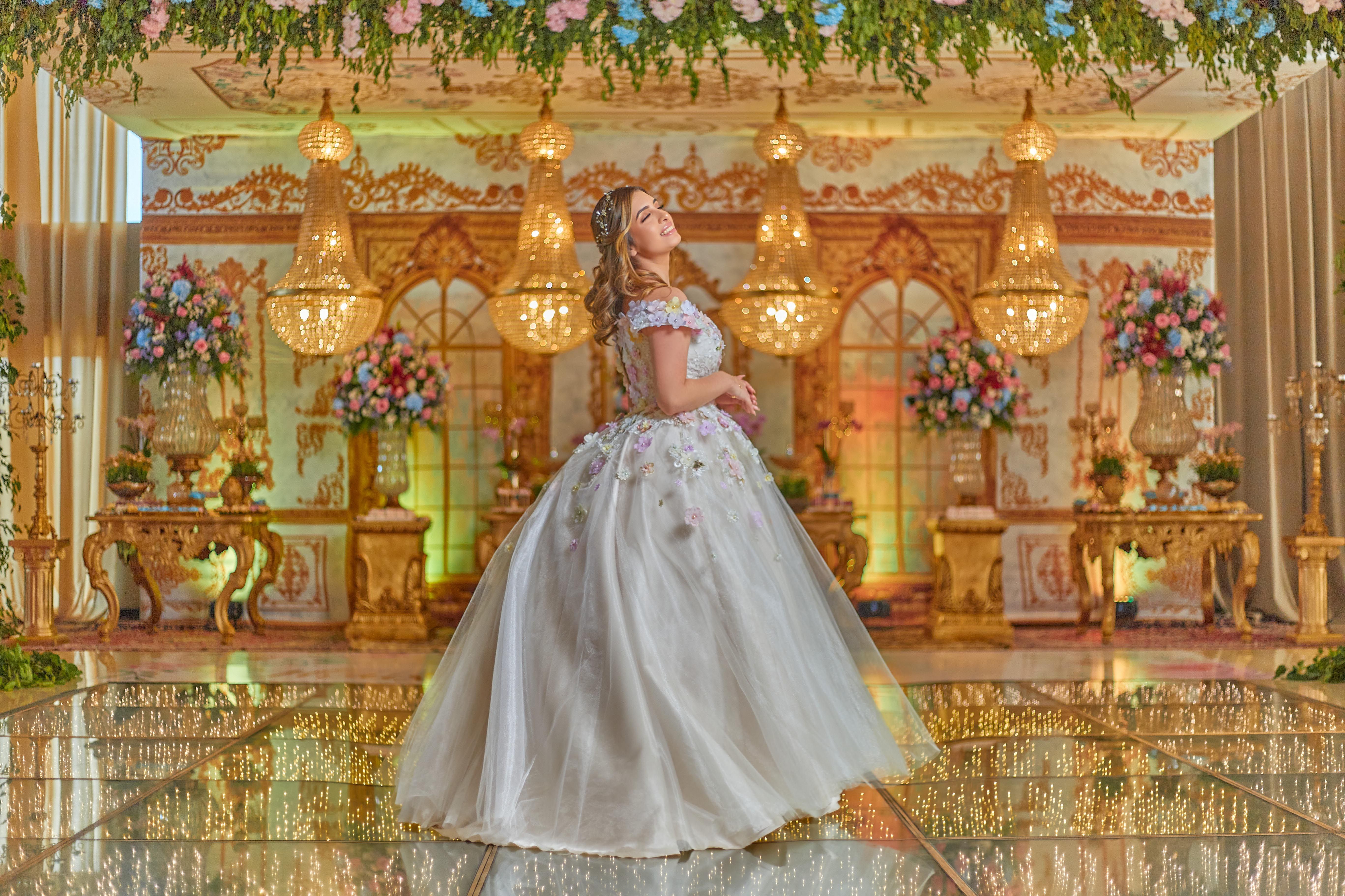 A young woman in a white gown poses elegantly in a luxurious ballroom with chandeliers and floral arrangements.