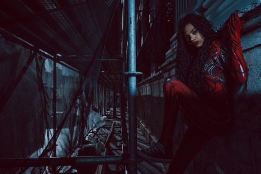 A woman in a red costume sits on scaffolding in an urban setting, creating a moody atmosphere.