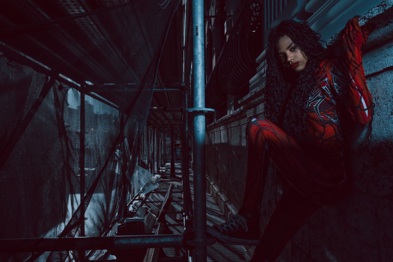 a woman in a red costume sits on scaffolding in an urban setting creat