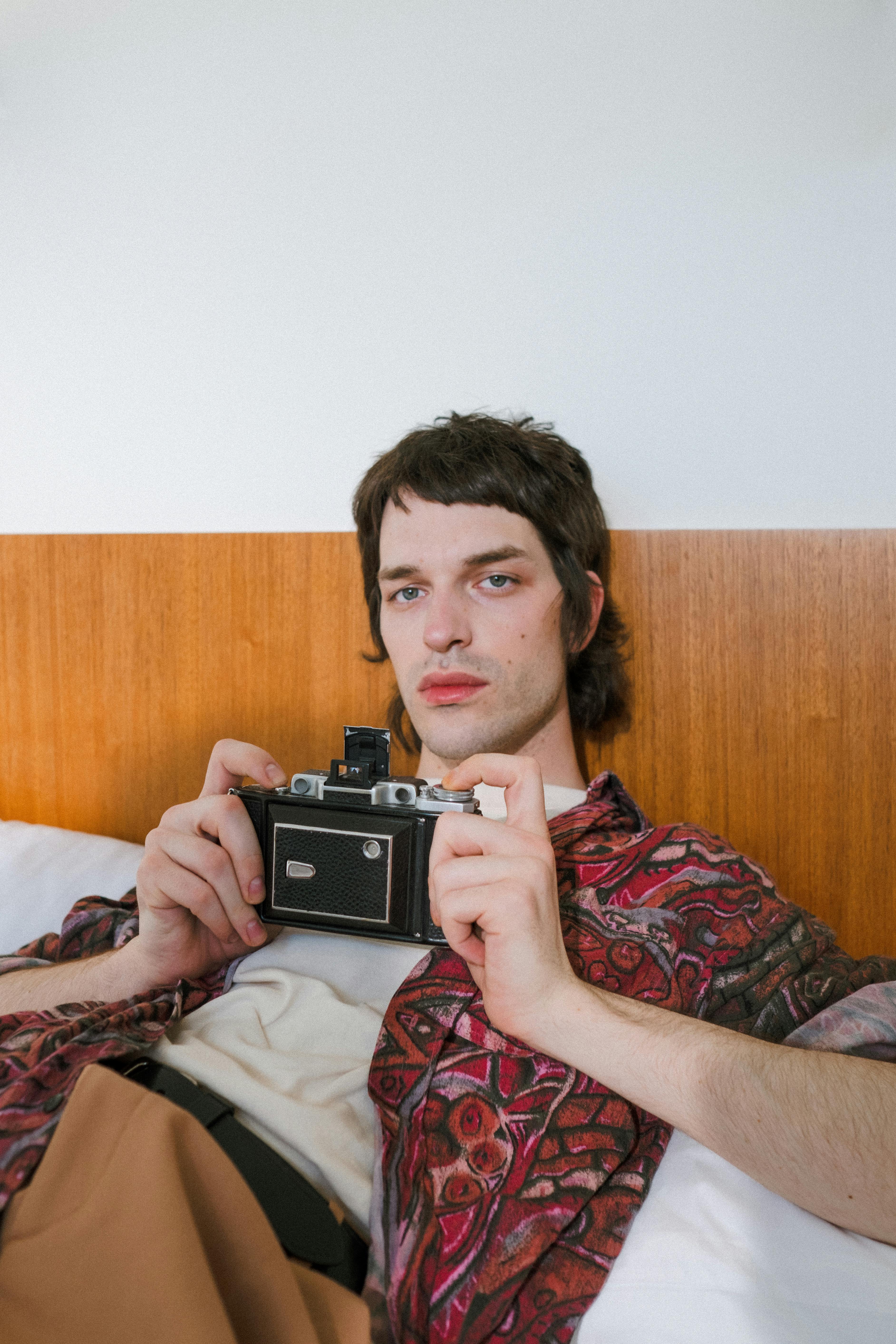 a shot of a male laying on bed holding an analog camera and looking at camera