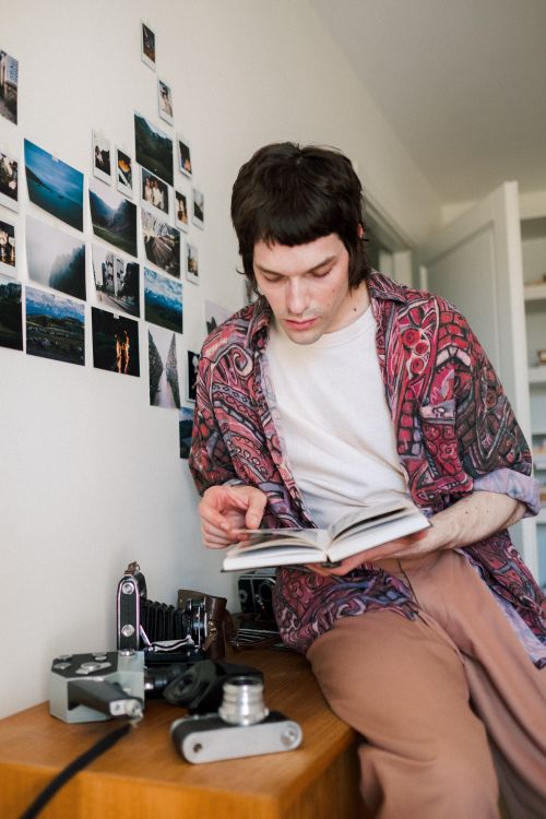 Free A young man in a patterned shirt reading a book indoors, surrounded by vintage cameras. Stock Photo