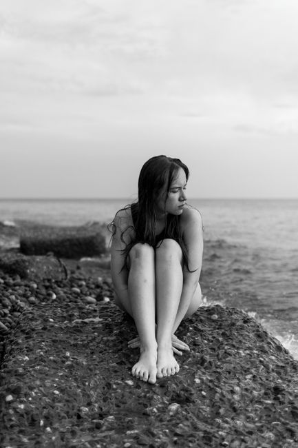 A thoughtful young woman sitting on a rock by the sea in a black and white setting.