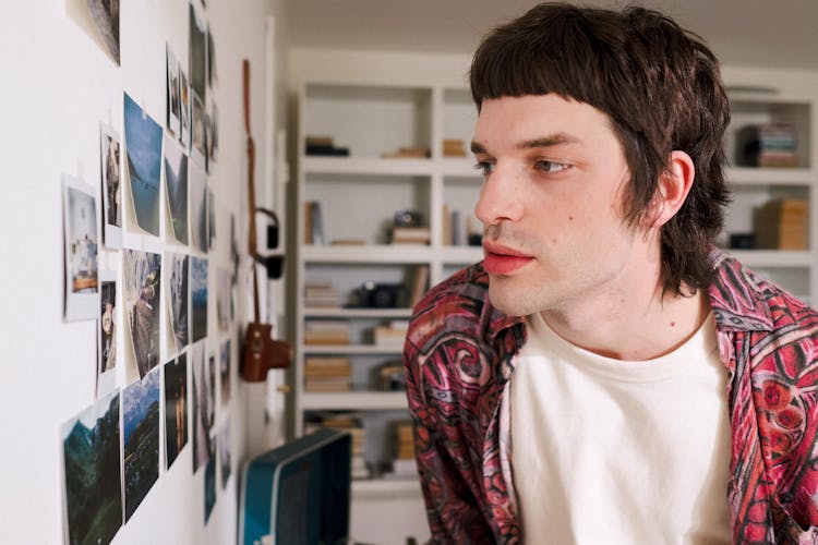 Young Man Looking At Photos Pinned To Wall In Room