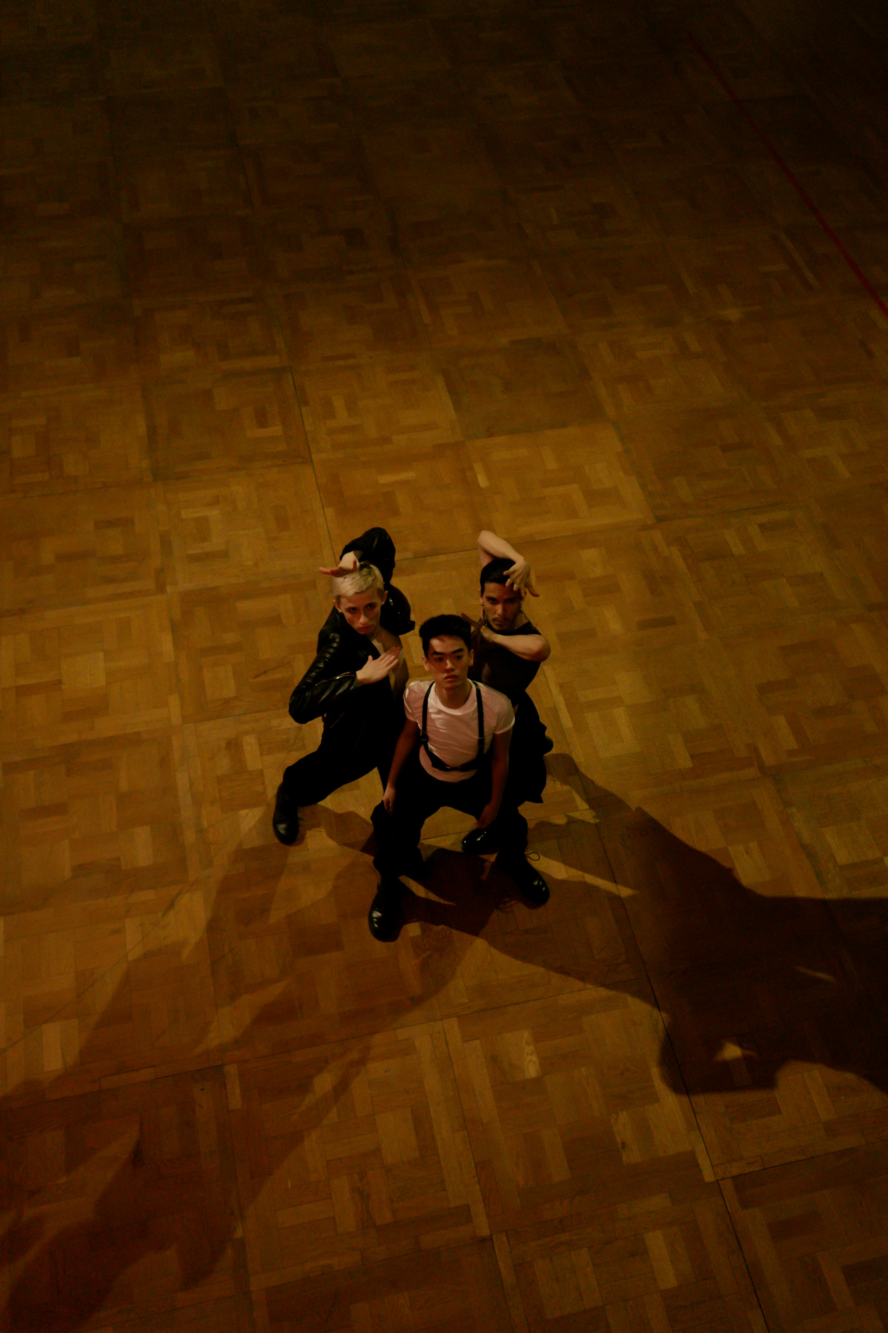 Three adult performers engage in a dynamic dance routine on a wooden floor, captured from above.