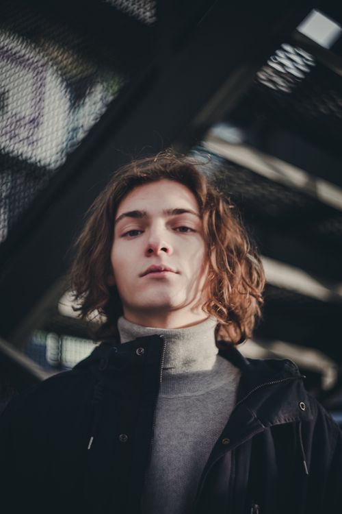 Free Portrait of a confident young man with long hair in casual attire in an outdoor urban setting. Stock Photo