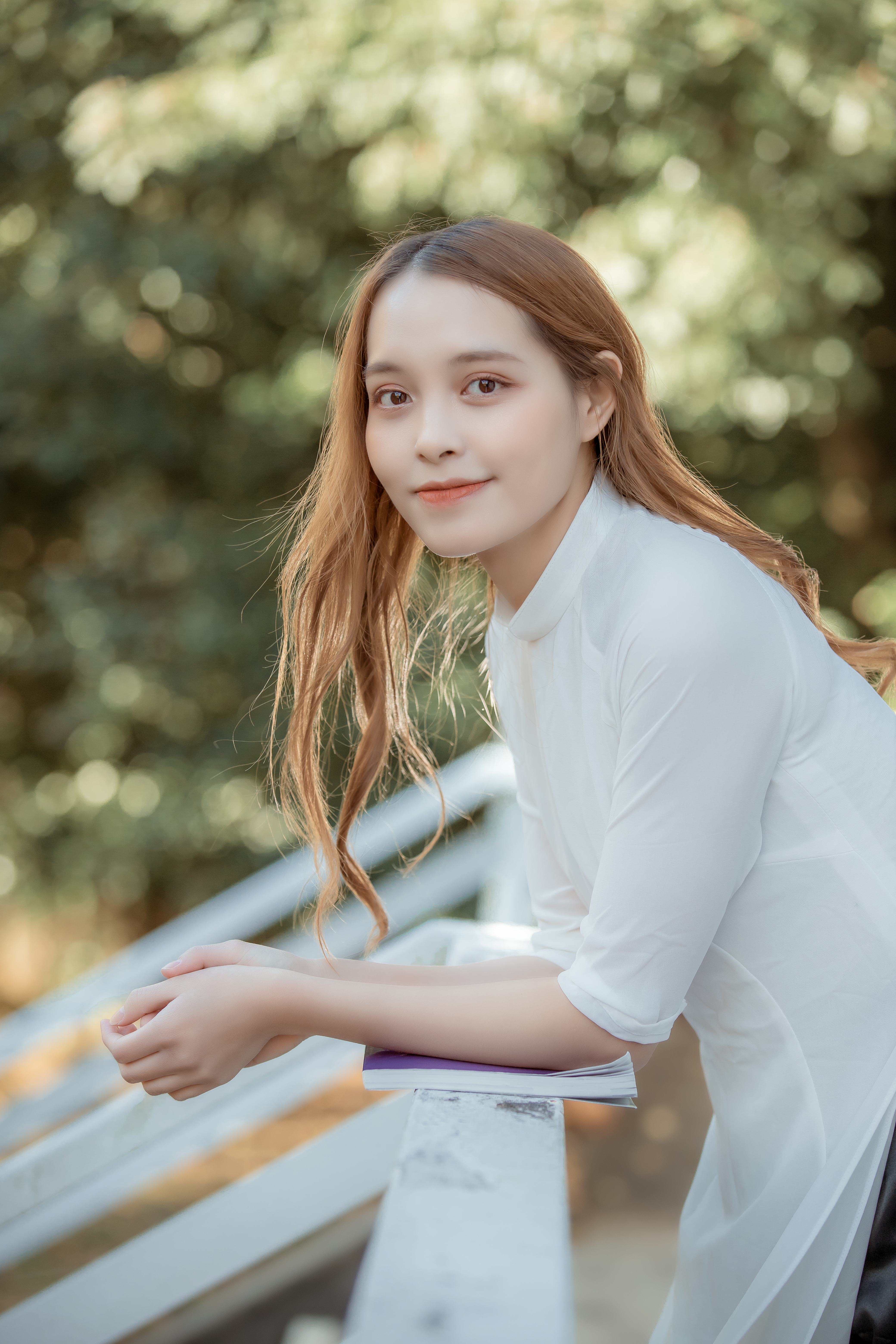 A young woman in a white shirt leans casually on a rail in a sunny outdoor setting.