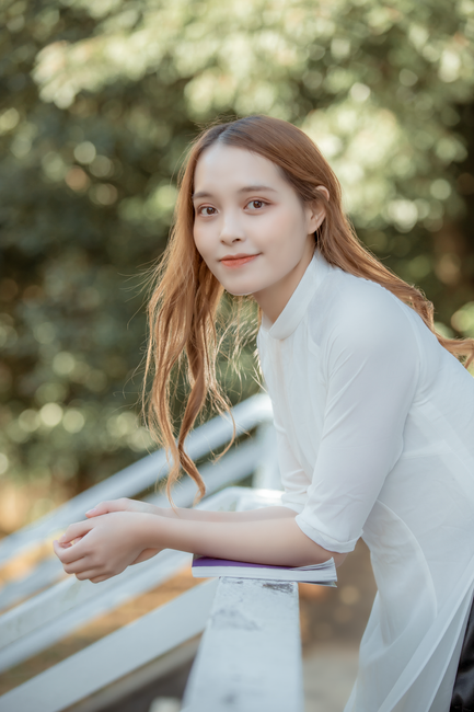 A young woman in a white shirt leans casually on a rail in a sunny outdoor setting.