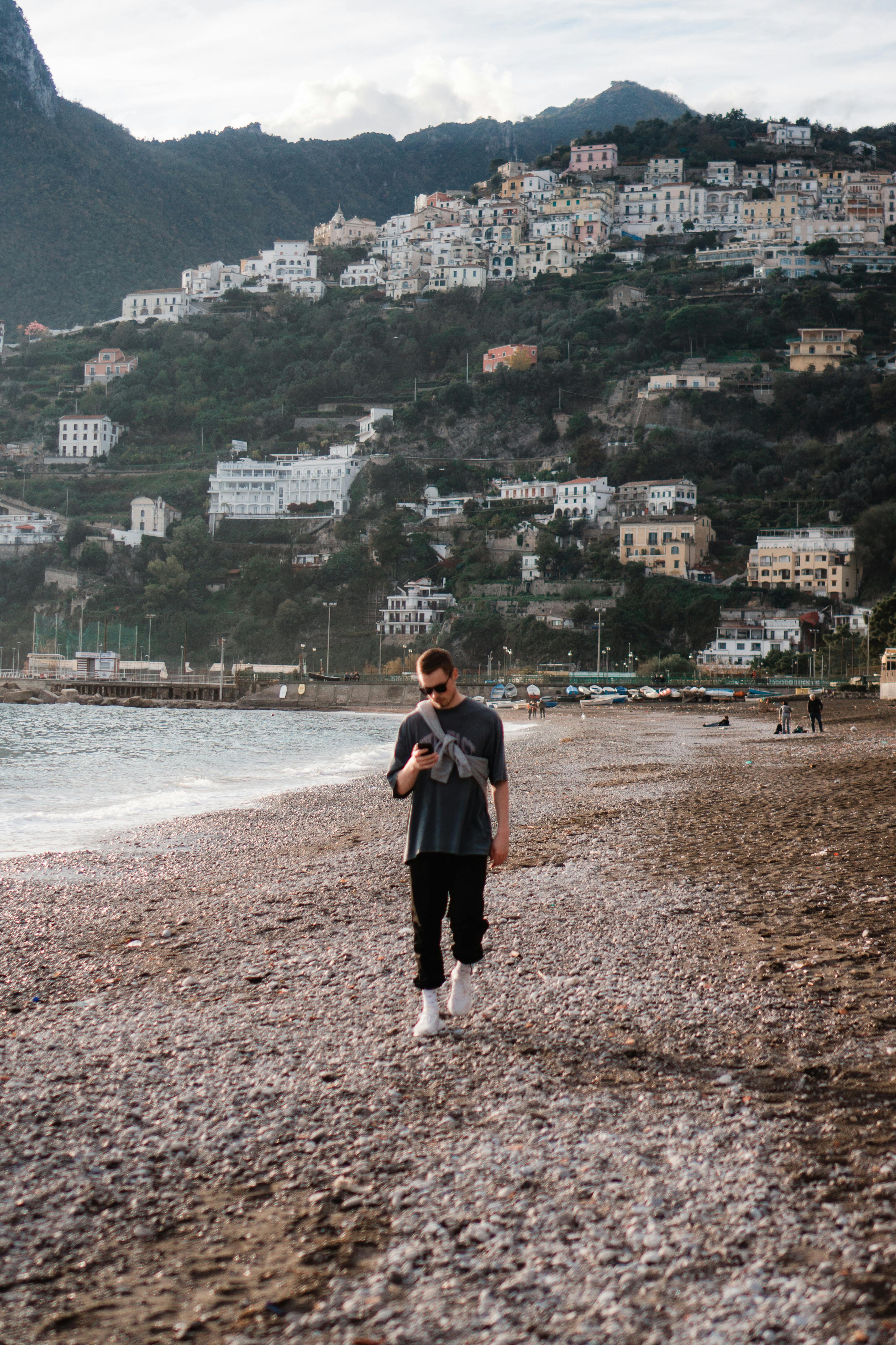 A man in streetwear walks on a pebble beach with scenic Italian hillside town in the background.