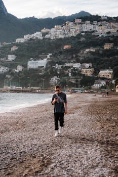 A man in streetwear walks on a pebble beach with scenic Italian hillside town in the background.