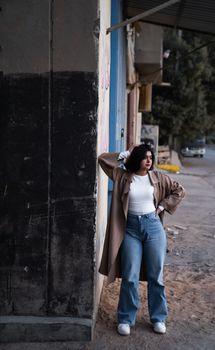 Confident young woman poses stylishly on an urban street, wearing a brown coat and denim jeans.