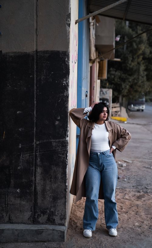 Free Confident young woman poses stylishly on an urban street, wearing a brown coat and denim jeans. Stock Photo